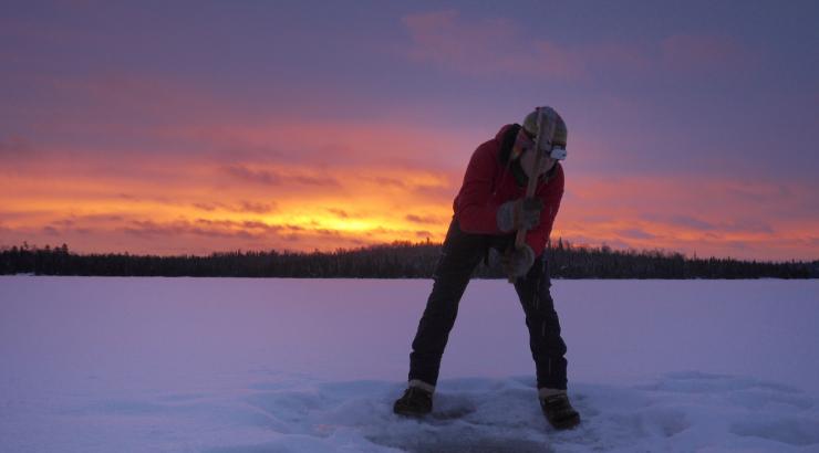 Photo of Amy Freeman digging an ice hole on a frozen lake next to a Boundary Waters sunset