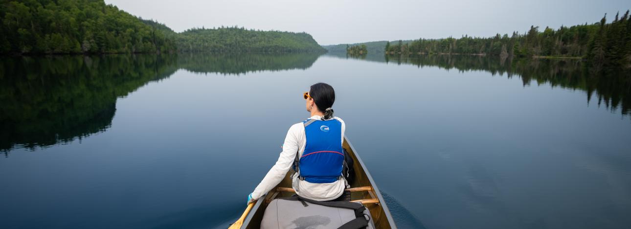 View from behind a woman canoeing on wilderness lake that is a calm and still with palisades in the distance. 